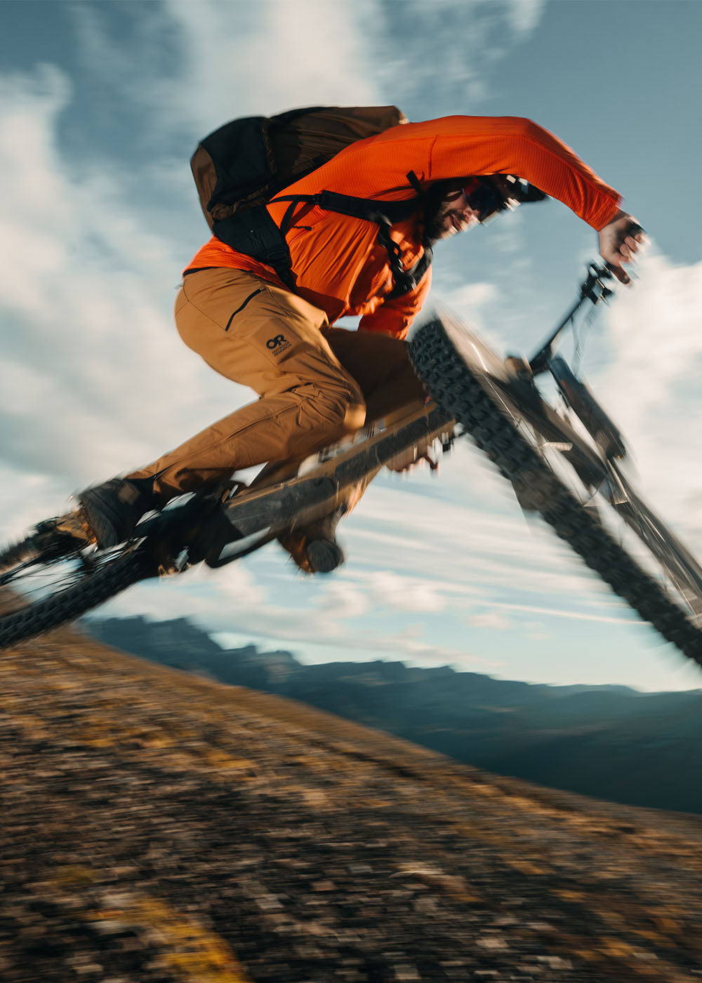 Male in the air on his mountain bike in the mountains wearing Outdoor Research Men's Deviator Hoodie Lingonberry and Men's Freewheel MTB Ride Pants Bronze. 