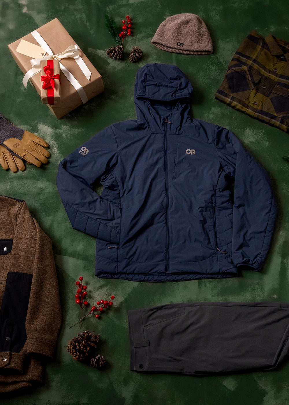 Mens Outdoor Research sportswear and outerwear lies on a dark green drop cloth with brown gift boxes, holly, and pine cones.