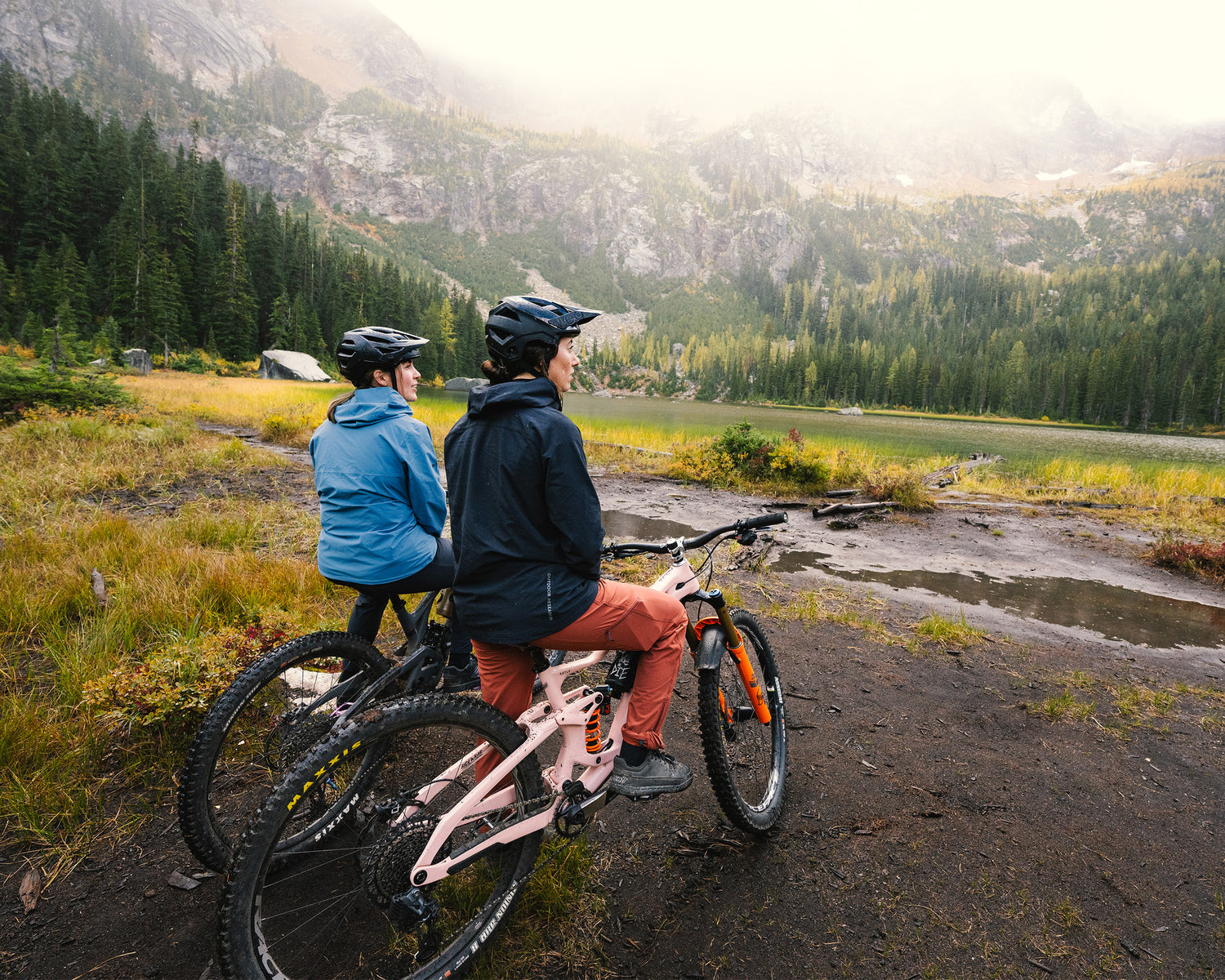 Two mountain bikers take in a view of the mountains while sitting on their bikes on a wet, muddy day.
