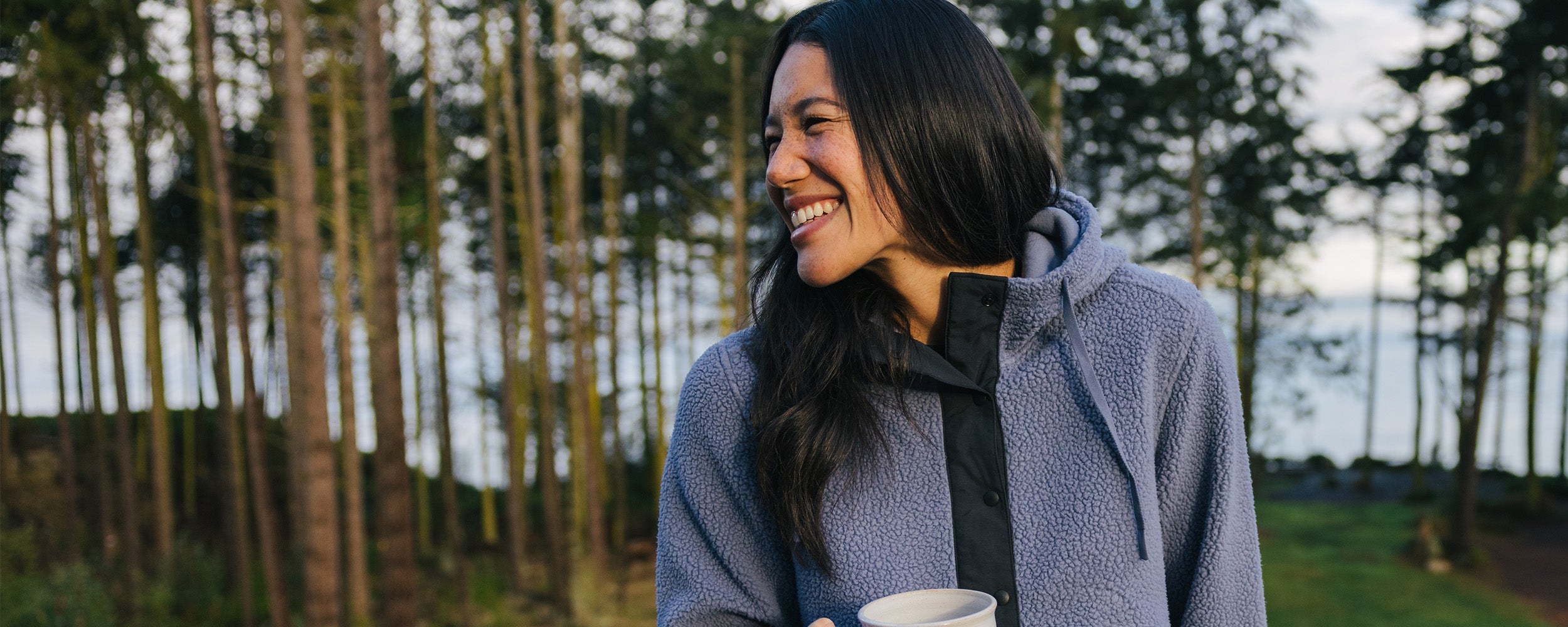 A female laughing and leaning against the deck with a cup of coffee wearing Outdoor Research Women's Grayland Fleece Pullover Hoodie Summit.