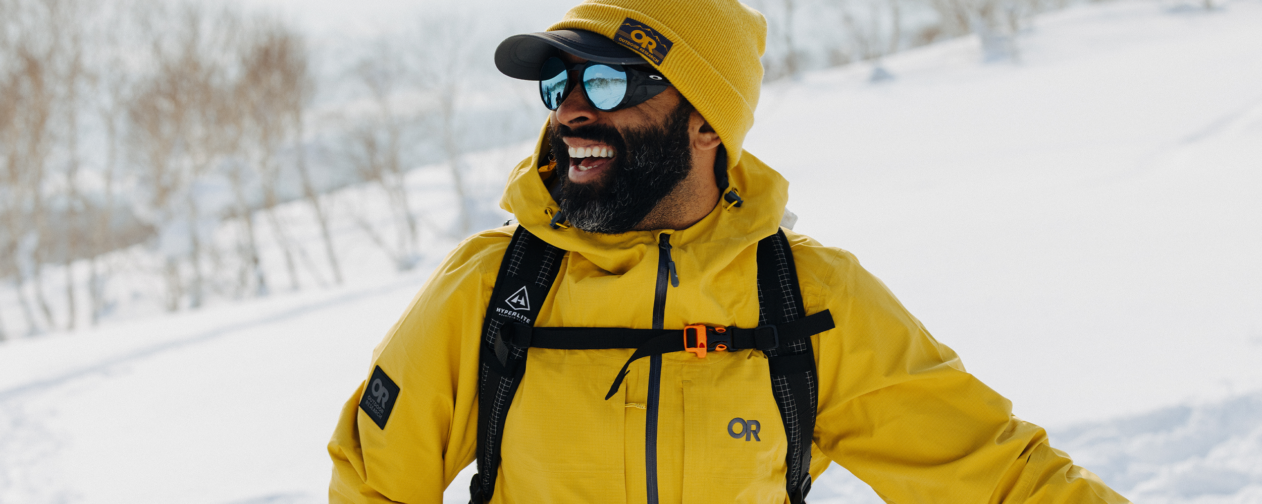 Male in the snow wearing Outdoor Research Men's Carbide Jacket Amber and Juneau Beanie Amber. 