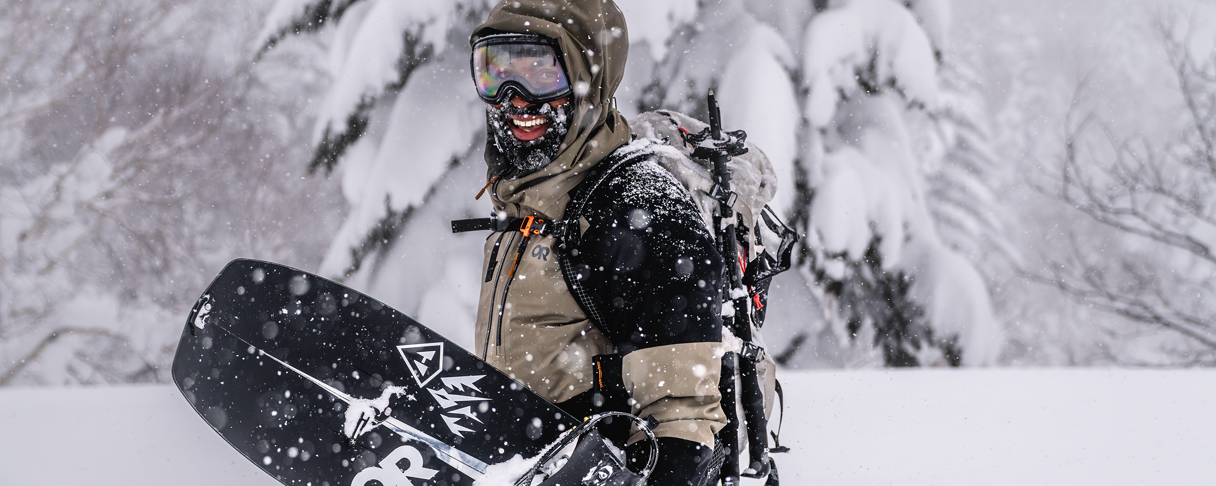 Male hiking through the snow with snowboard wearing Outdoor Research Men's Hemispheres Team GORE-TEX 3l Jacket Gravel/Black. 