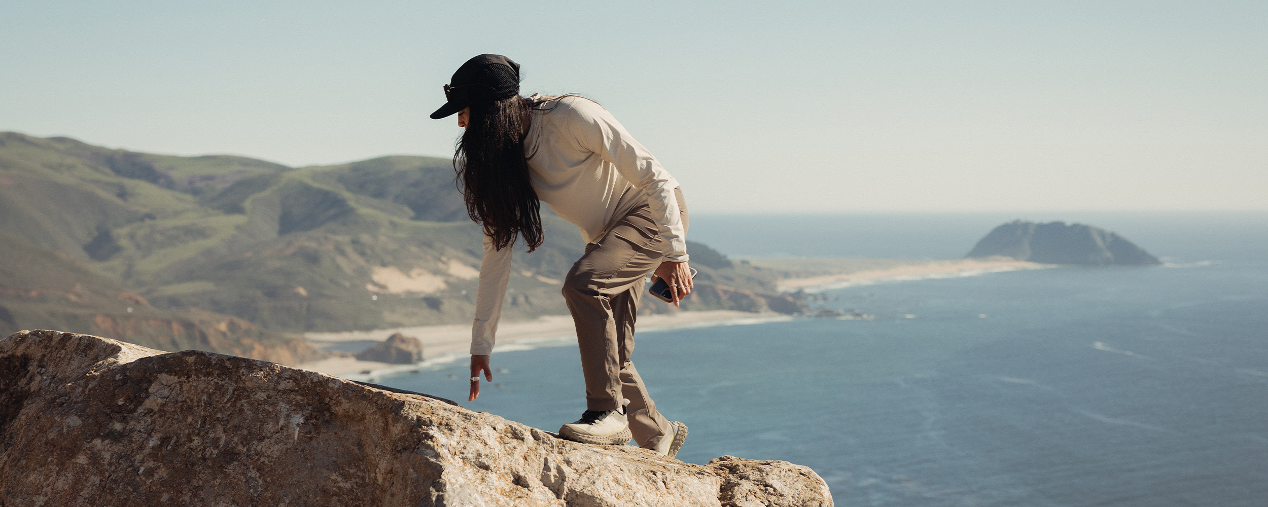 Woman wearing Outdoor Research Women's Vigor LT Hoodie Oyster on the rocks by the water. 