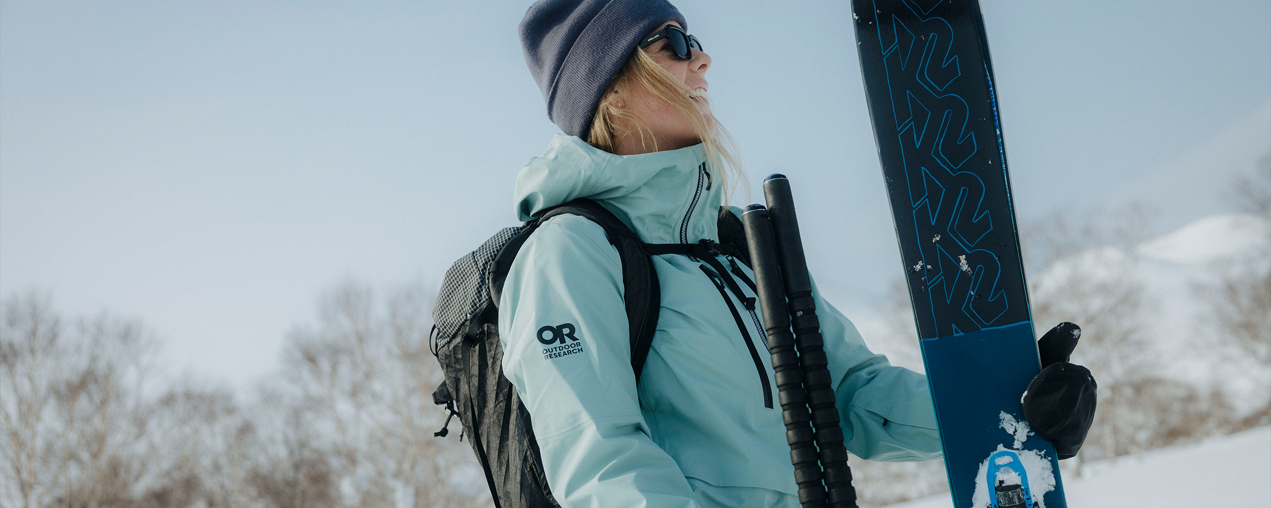 Female holding skis wearing Outdoor Research Skytour LT Jacket Thyme. 