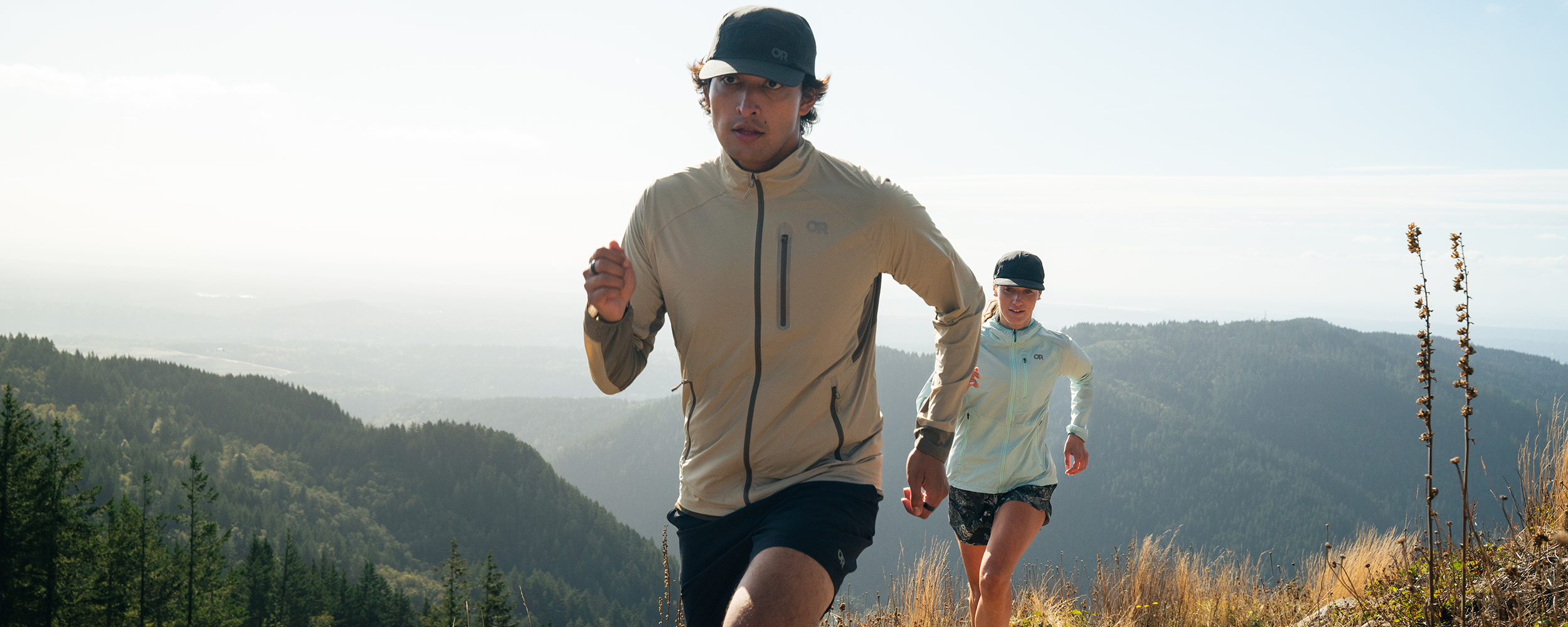 Two runners jog across a bridge along the water wearing Outdoor Research Sportwear. 