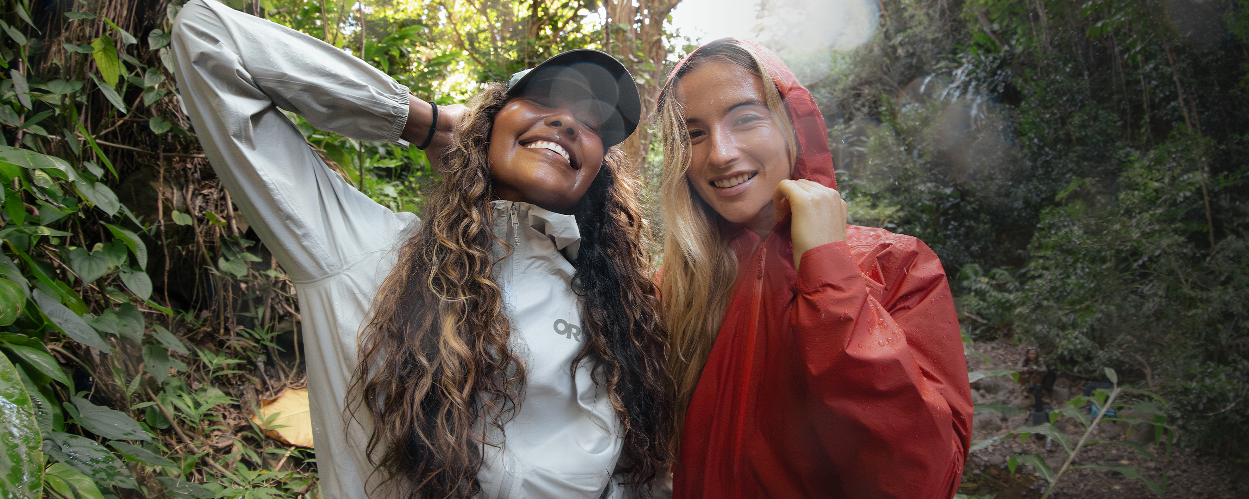 Two females smiling in the rain wearing Outdoor Research Women's Helium UL Jackets Madrone Red and Oyster. 