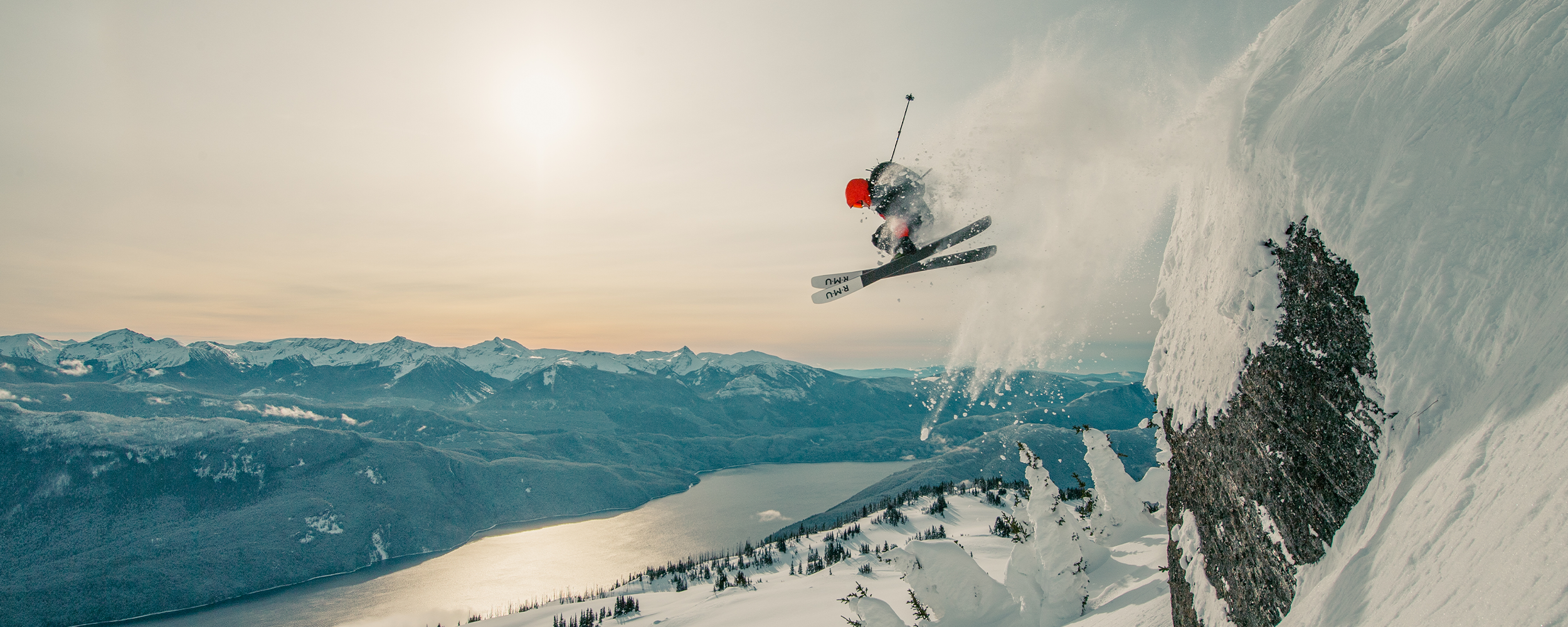 Skier going off a big jump overlooking the water wearing Outdoor Research Men's Powderverse Jacket Lingonberry/Black.
