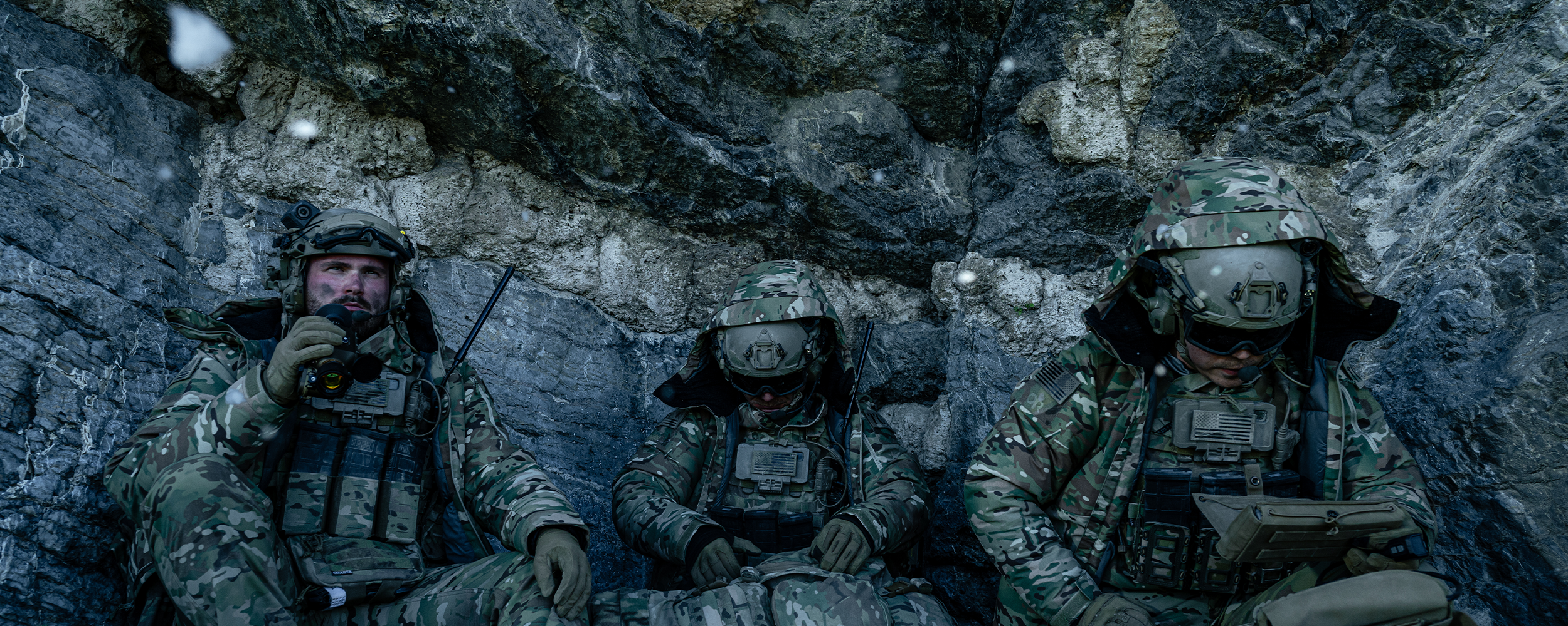 A group of men wear tactical camouflage while leaning up against a rock wall.