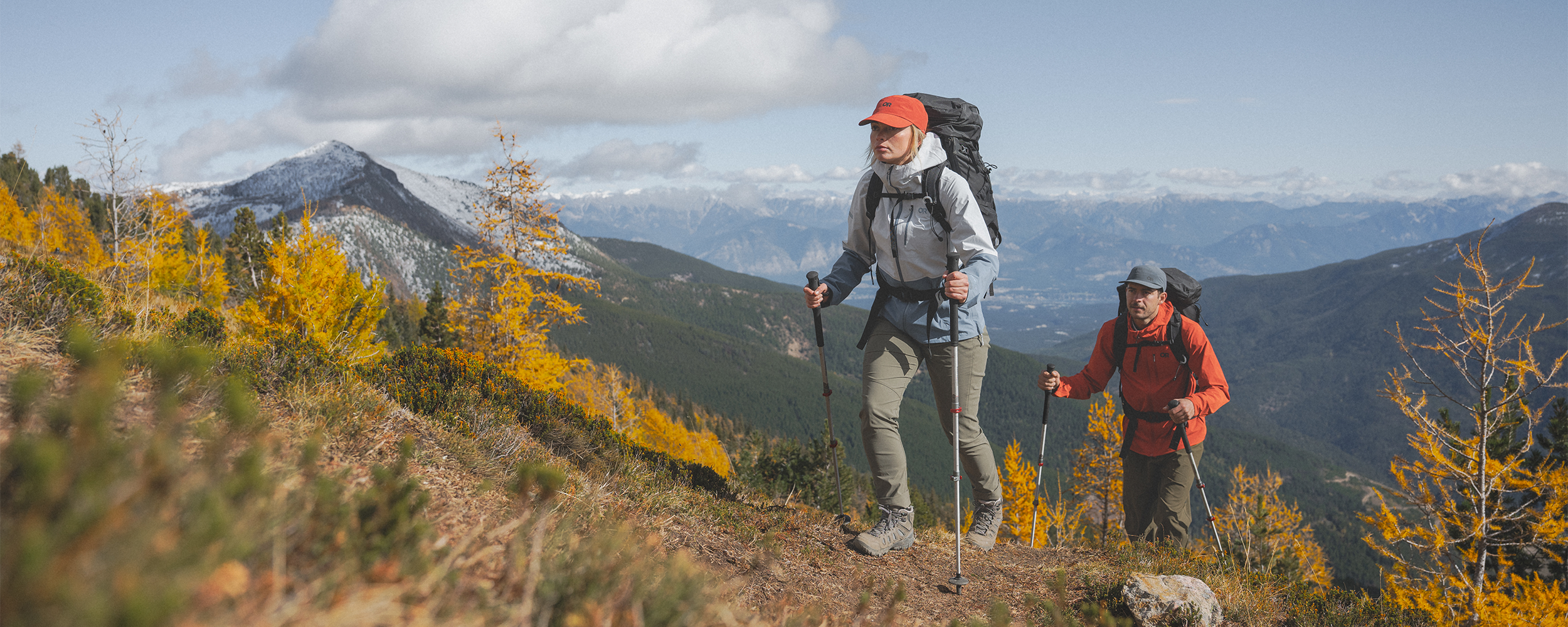 Two hikers on a sunny day wearing Outdoor Research Women's Aspire 3L Jacket Titanium/Ascent Blue, Women's timberline Pant Ranger Green, Mens Ferrosi Hoodie Cardinal. 