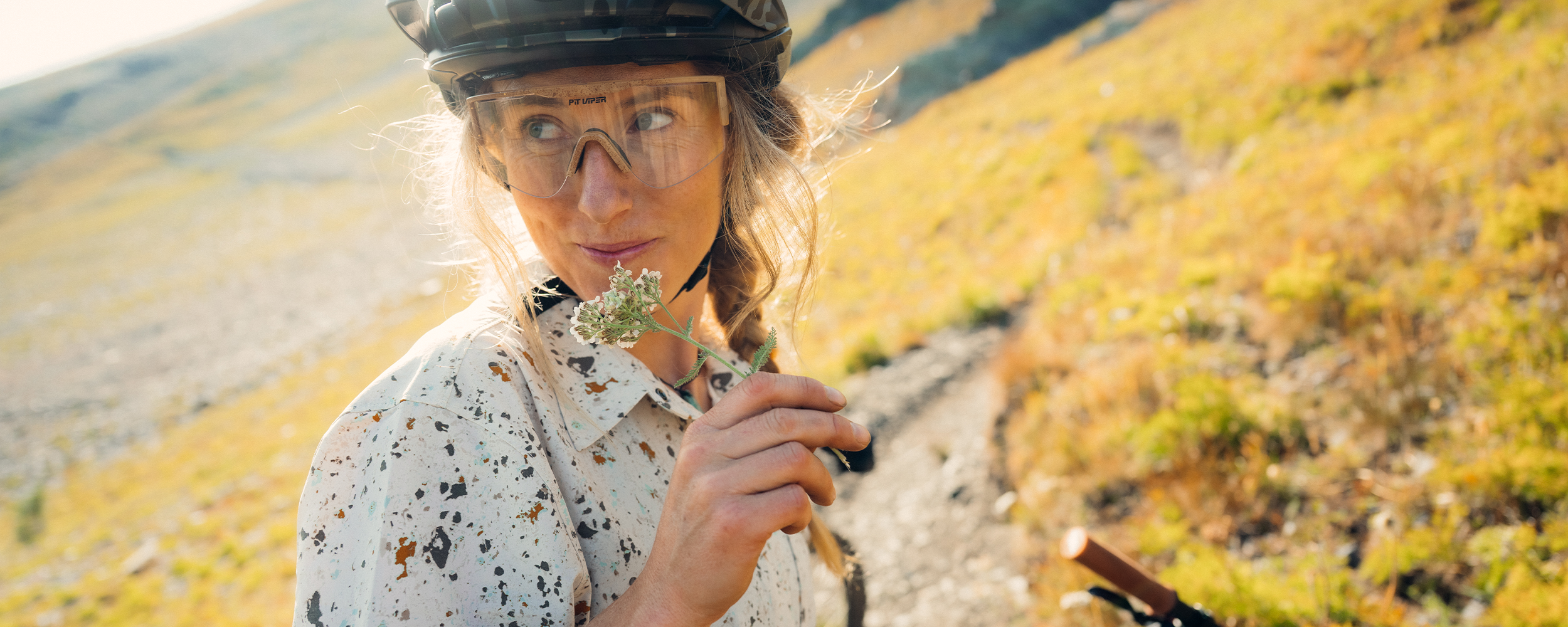 Female holding flowers in her hand in the sun wearing Outdoor Research Women's Astroman Short Sleeve Sun Shirt Gravel Creekside Print.