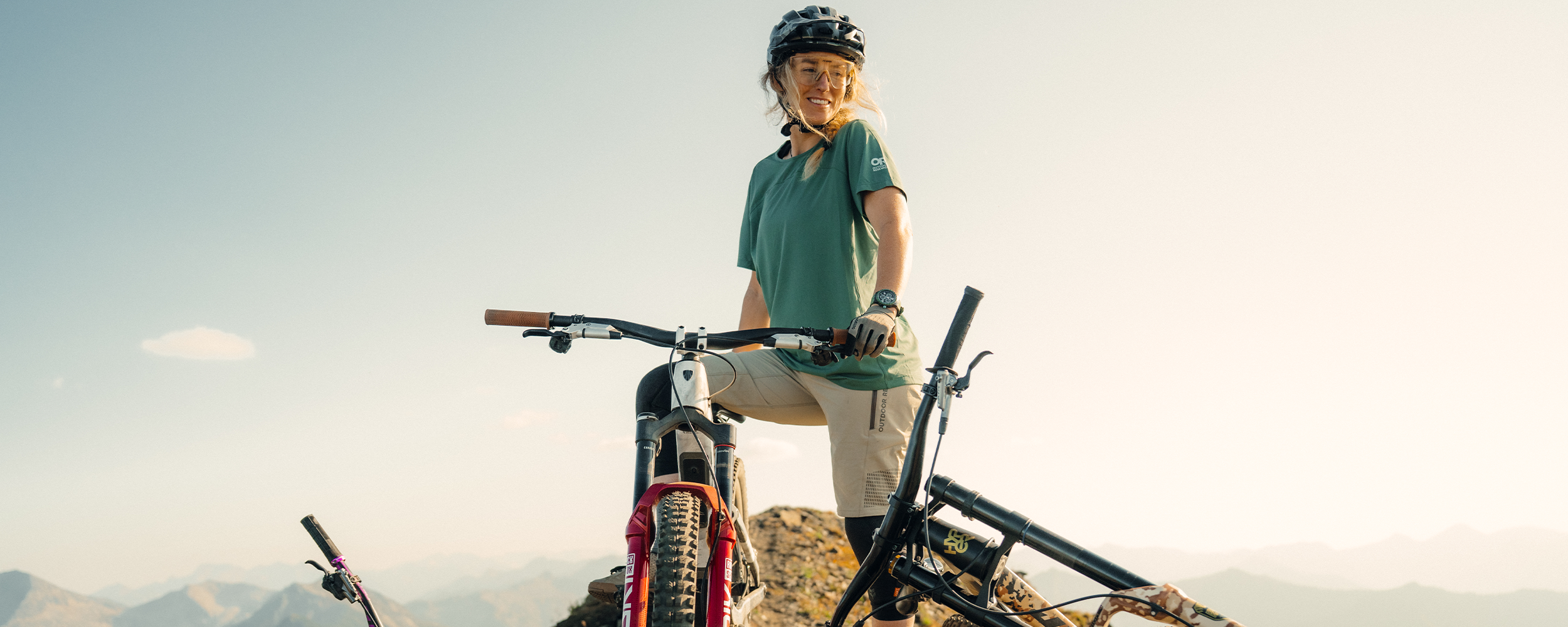 Female standing at the top of the hill with her bike wearing Outdoor Research Women's Freewheel MTB Ride Short and Women's Freewheel MTB Short Sleeve Jersey Malachite. 