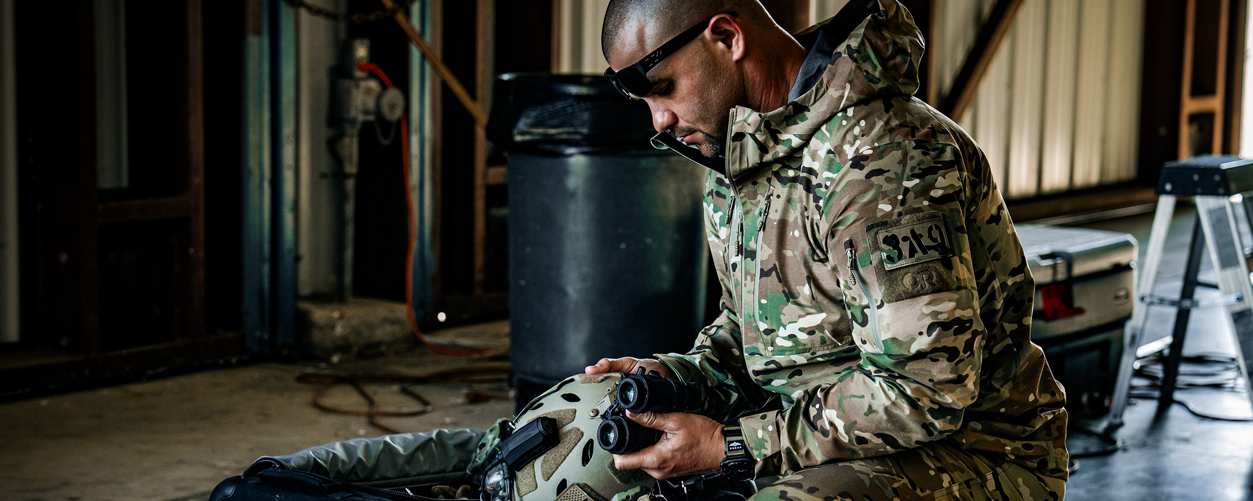 Two men in tactical gear lay atop a rock looking out above a lake and valley.