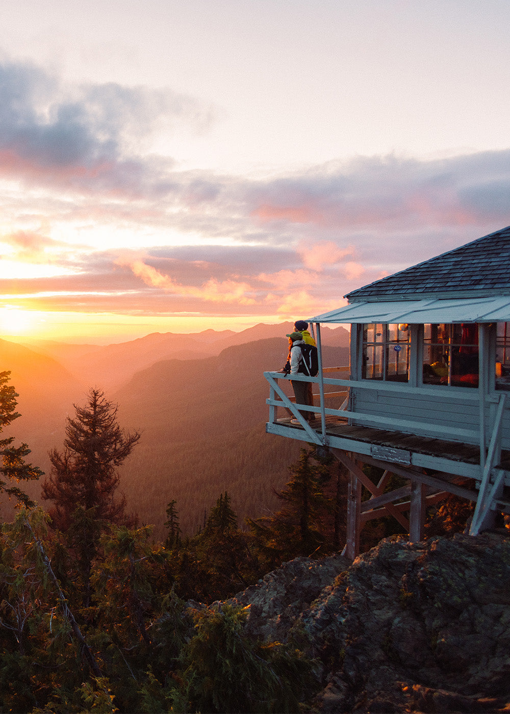Two friends stand on a deck and watch the sunrise glow behind the mountains while wearing Outdoor Research Outerwear.