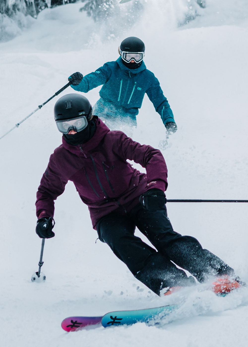 2 friends skiing down the mountain wearing Outdoor Research Women's Powderverse Jacket Winterberry and Aurora and Women's Powderverse Pants Black and Sable. 