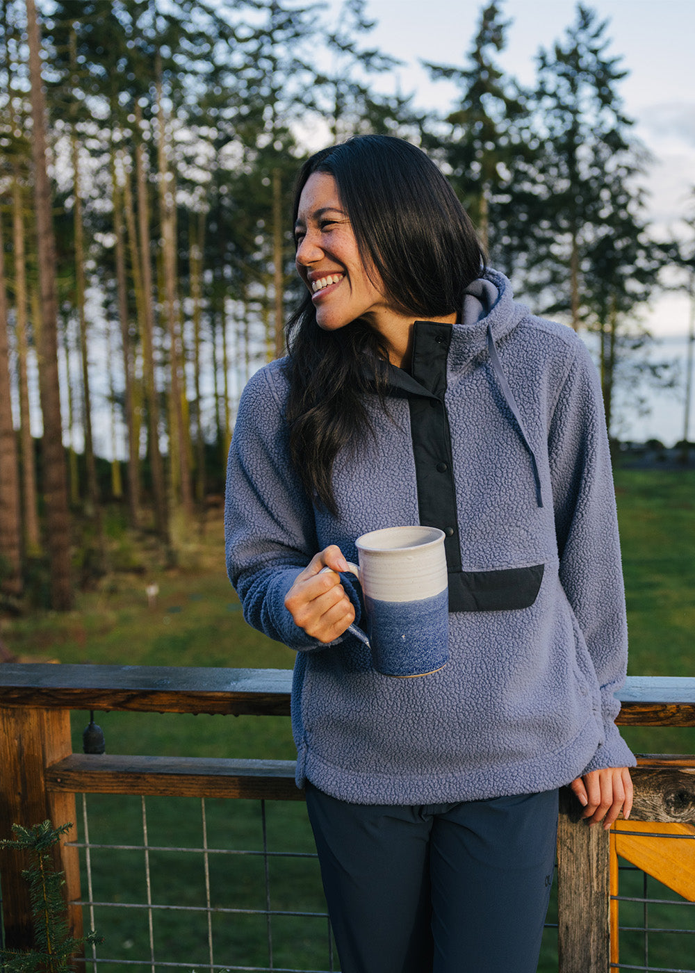 A female laughing and leaning against the deck with a cup of coffee wearing Outdoor Research Women's Grayland Fleece Pullover Hoodie Summit.