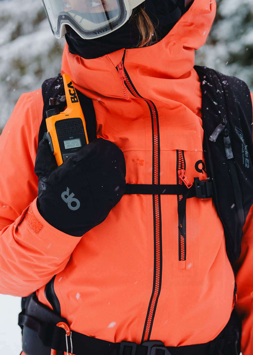 Person wearing an orange jacket and black gloves with a backpack in a snowy environment.
