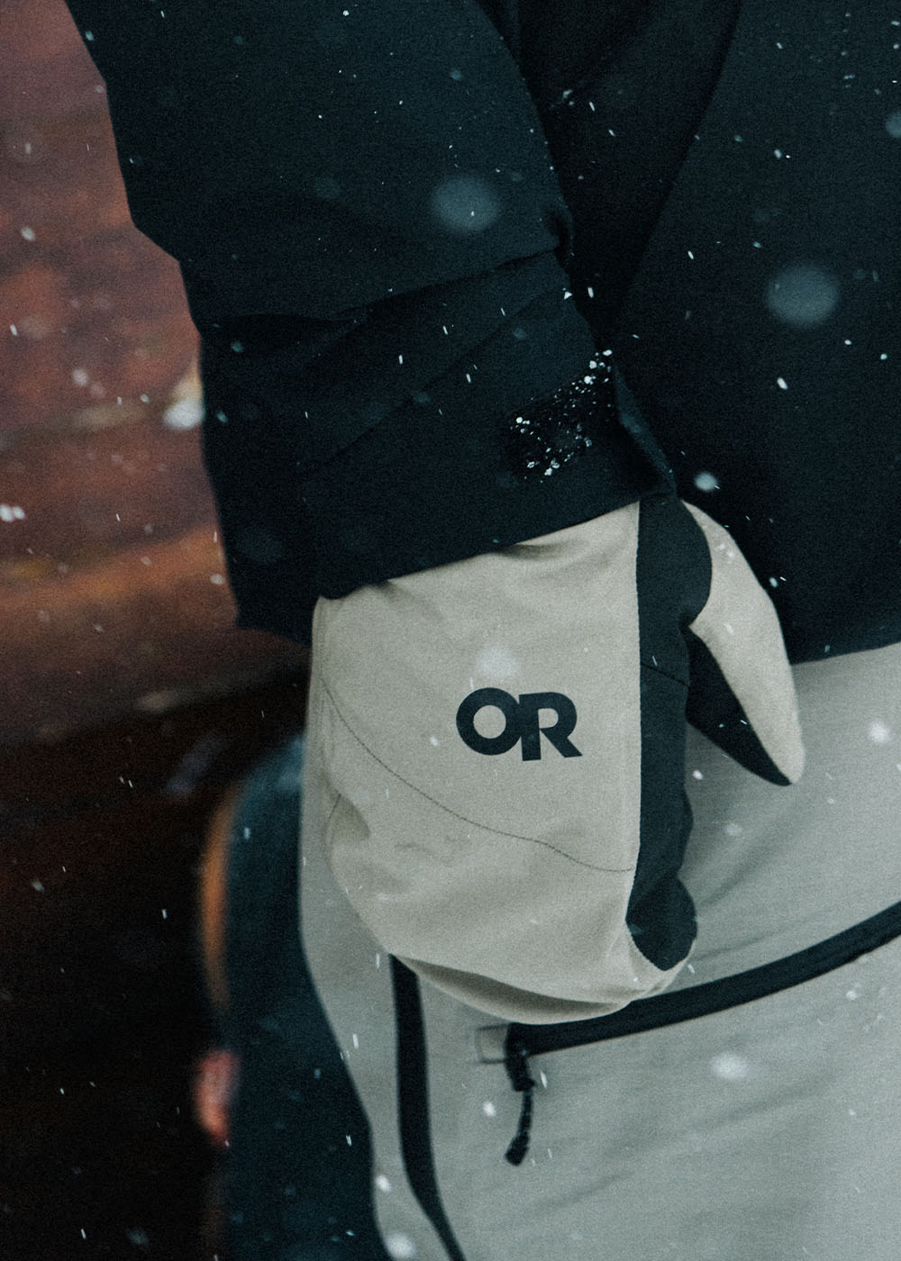 Male standing by the wall looking out at the snow wearing Outdoor Research Men's Powderverse Jacket Black and Hemispheres Team GORE-TEX 3L Mitts Sable. 
