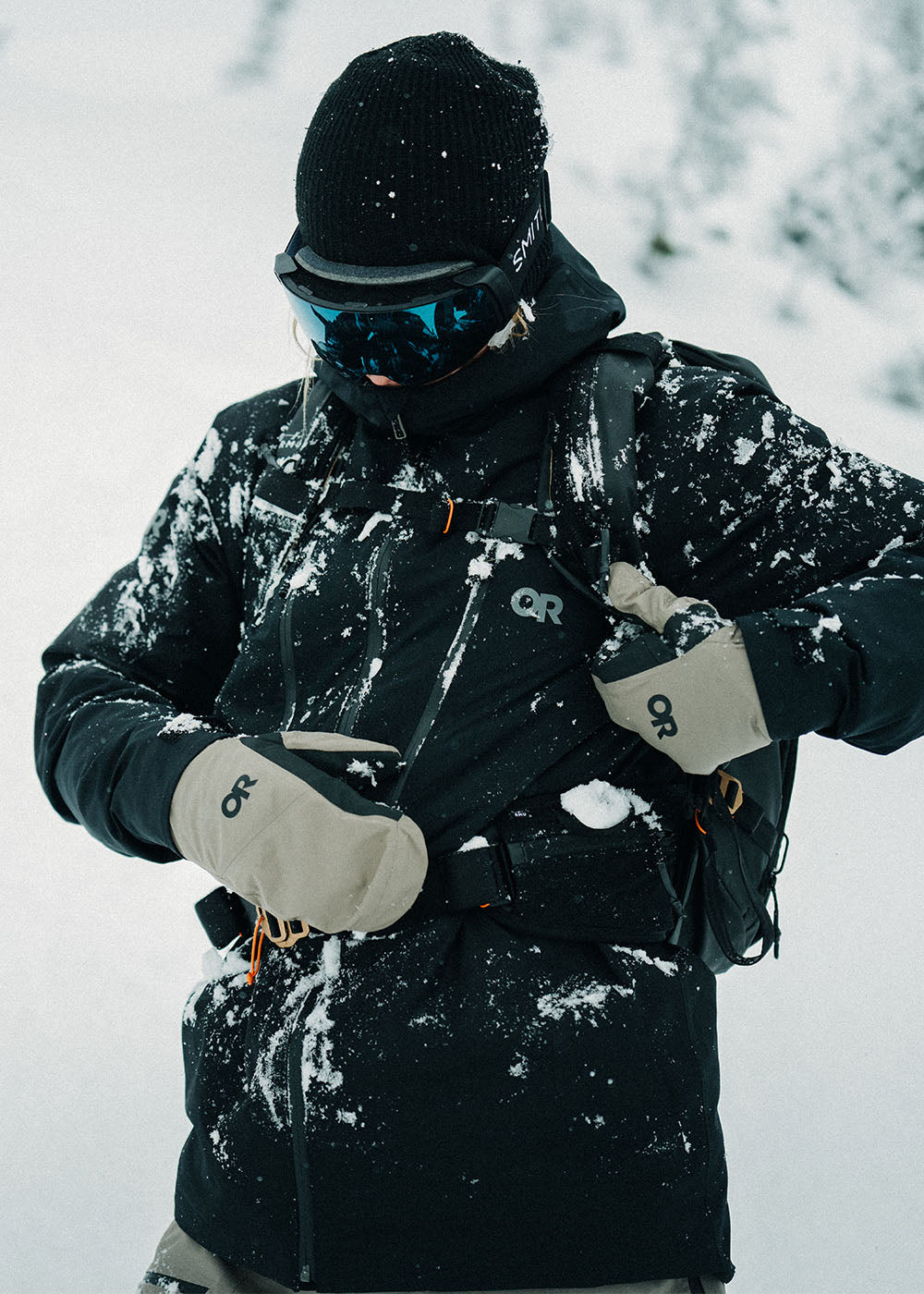A skier wearing the Outdoor Research Hemispheres Team GORE-TEX 3L Mitts in Sable checks their backpack while out on the mountain.