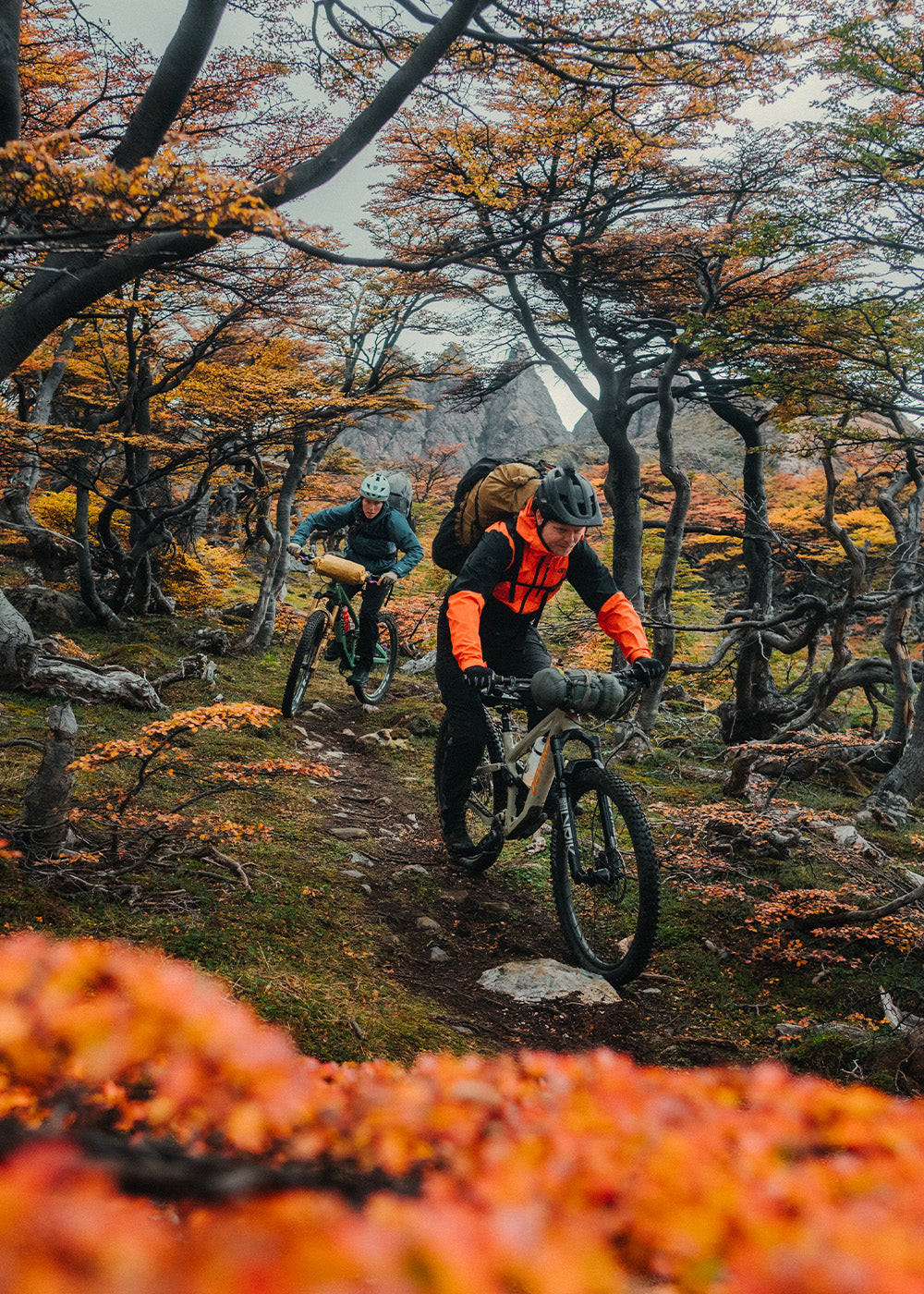 2 friends mountain biking through the trees wearing Outdoor Research Men's Freewheel MTB Stretch Rain Jacket Spice/Black and Women's Helium Rain Jacket Harbor. 