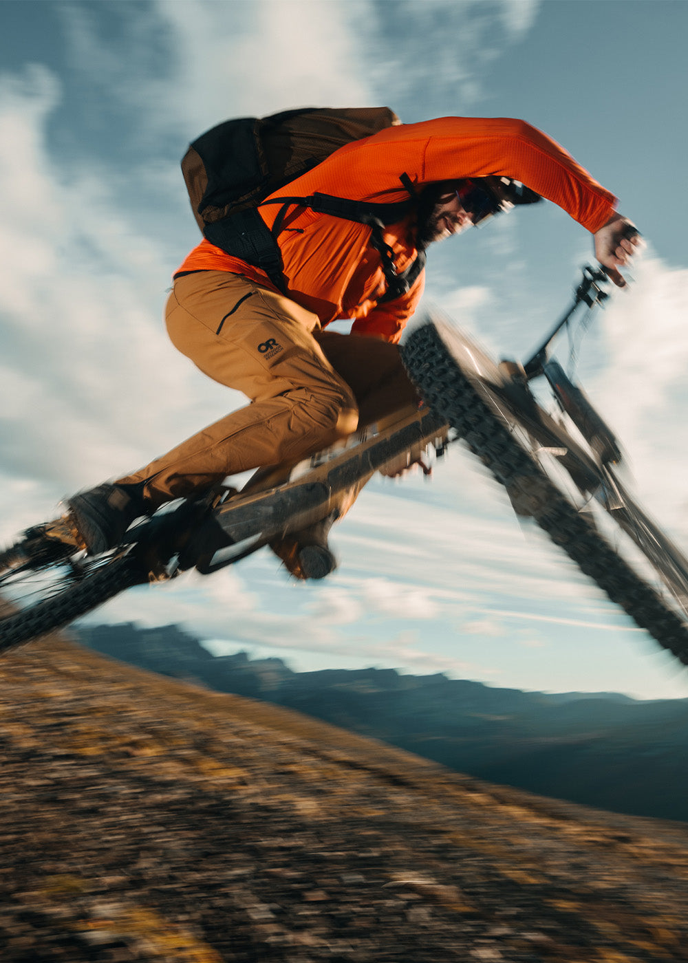 Male in the air on his mountain bike in the mountains wearing Outdoor Research Men's Deviator Hoodie Lingonberry and Men's Freewheel MTB Ride Pants Bronze. 