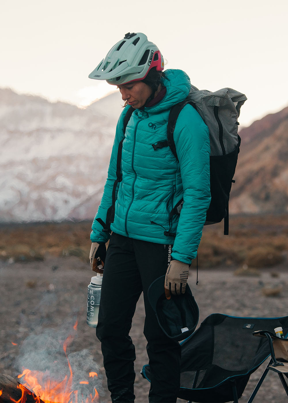Female standing around campfire with bike gear on wearing Outdoor Research Women's Super Strand XT Hoodie Tidepool and Freewheel MTB Leather Palm Bike Gloves Pro Khaki. 