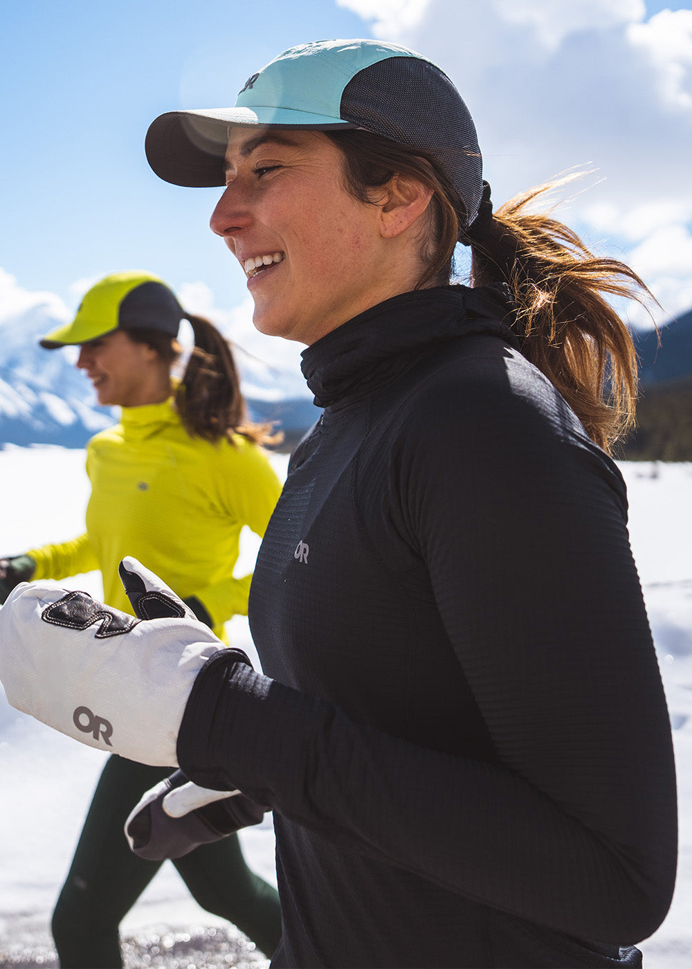 Two women run through snow wearing the Outdoor Research Vigor Grid Fleece Pullover Hoodie in Black.
