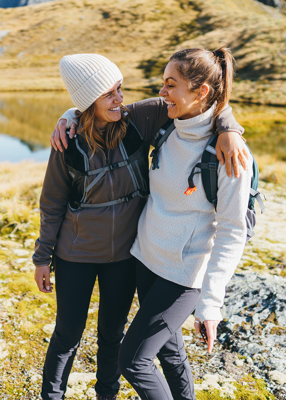 2 smiling female friends hugging in the mountains wearing Outdoor Research Women's Crescent Fleece Full Zip Hoodie Sable and Women's Trail Mix XT Fleece Cowl Oyster and Liftie VX Beanie Sand.