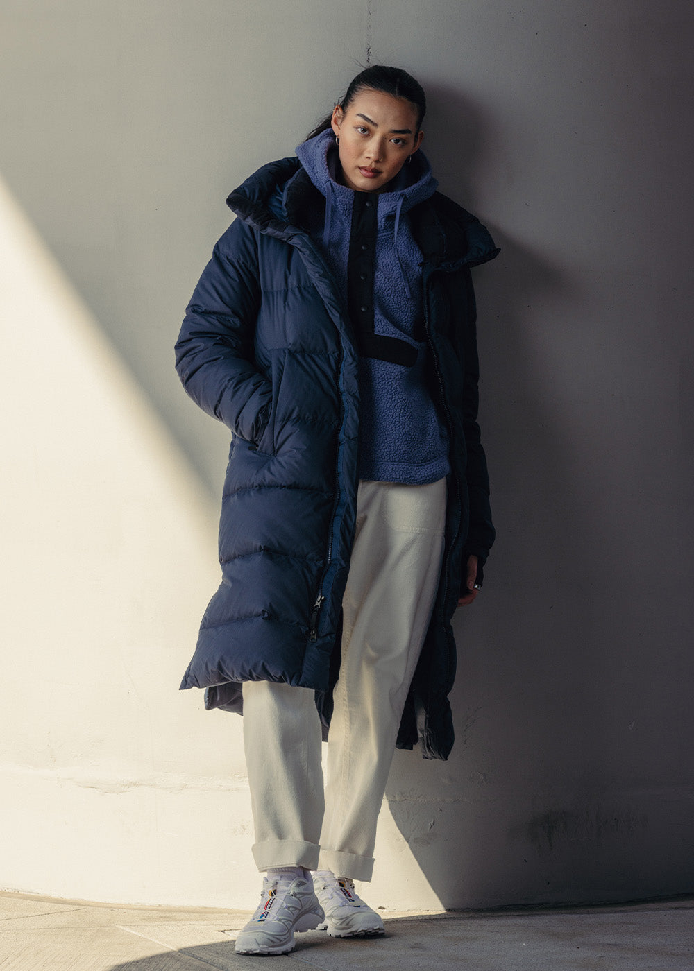 A woman stands by a wall wearing Outdoor Research Women's Coze Down Parka Atlantic and Women's Grayland Pullover Hoodie Summit. 