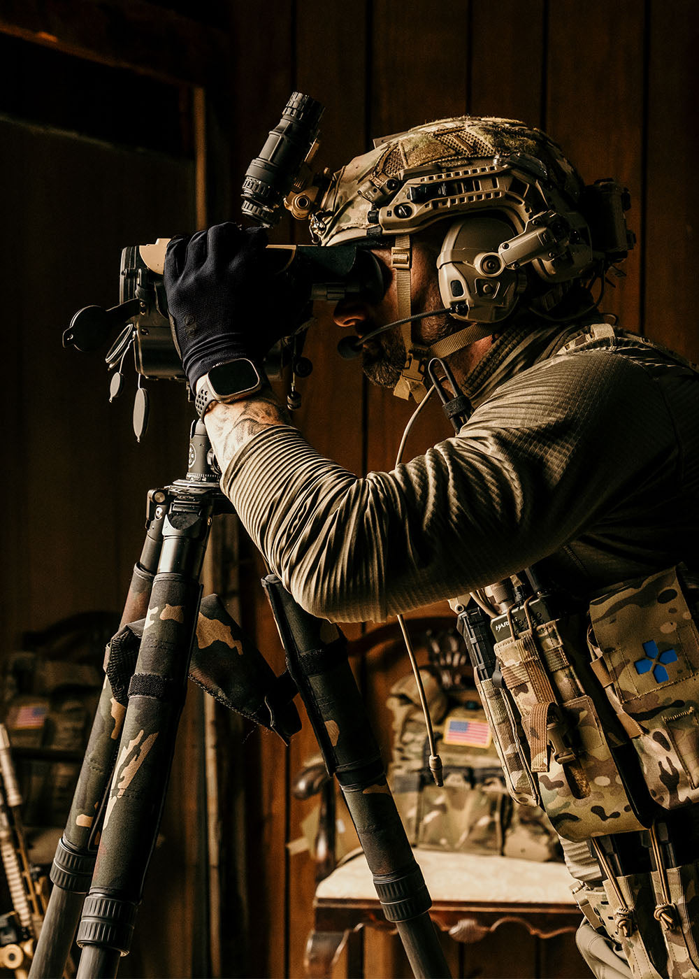 A person wearing Outdoor Research Tactical clothing looks through binoculars while standing near an open window.