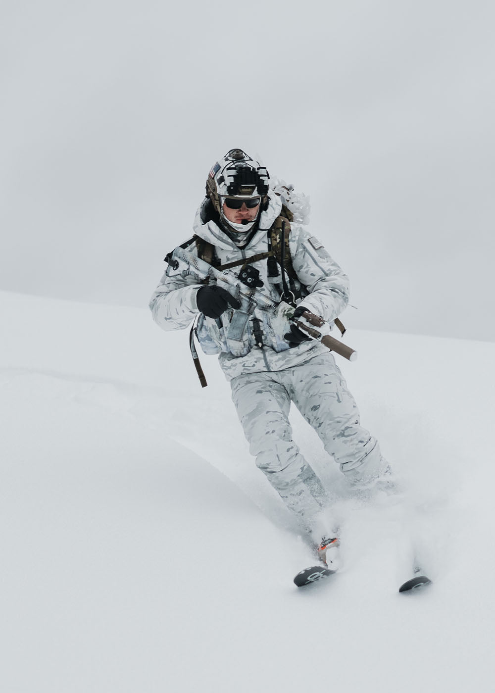 A man wearing alpine camouflaged tactical gear skis through snow while holding tactical equipment.