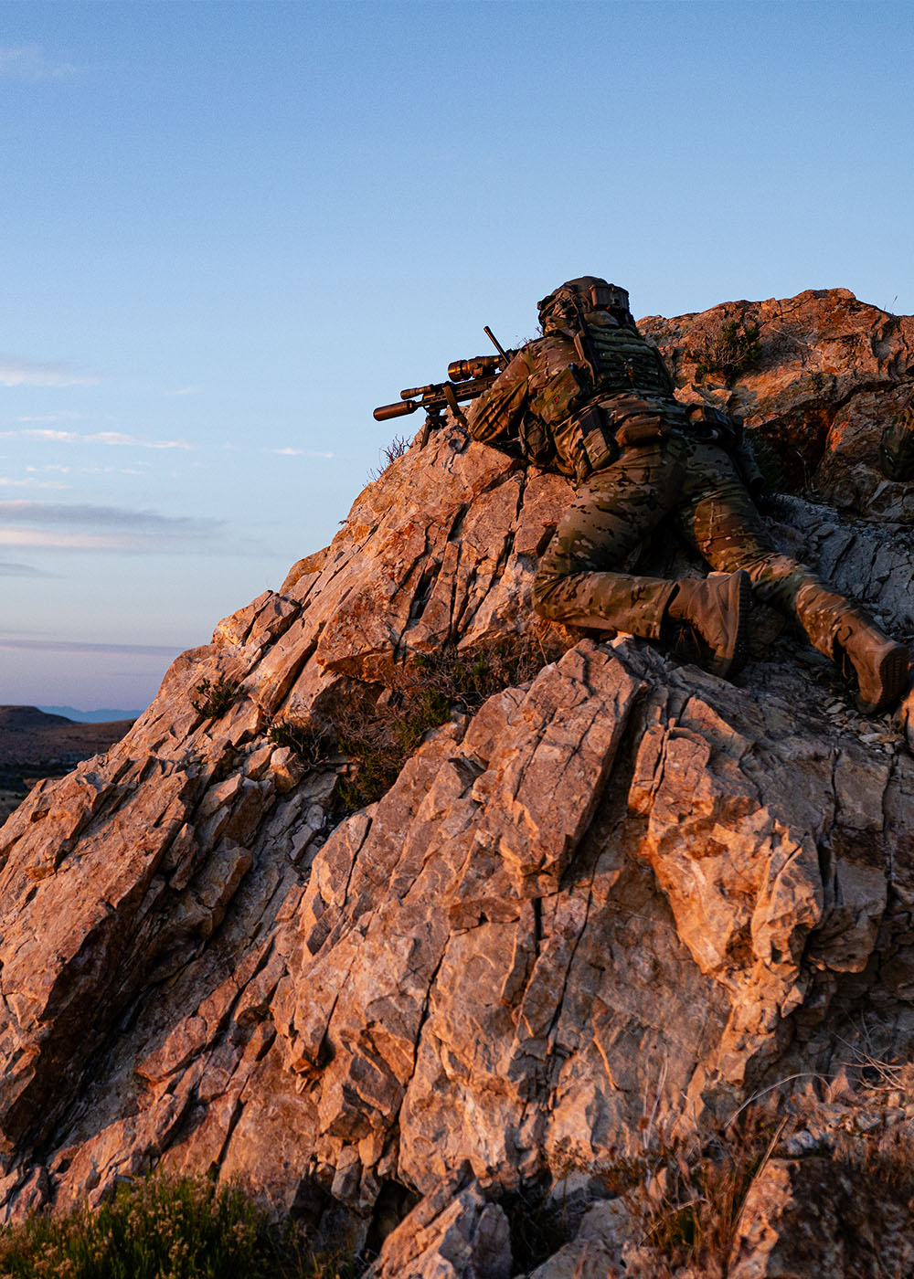 Two men in tactical gear lay atop a rock looking out above a lake and valley.