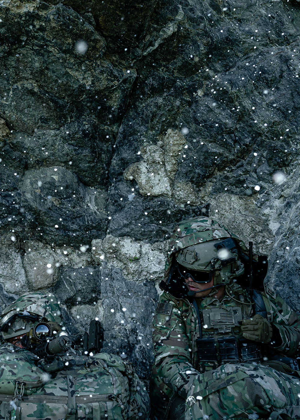 A group of men wear tactical camouflage while leaning up against a rock wall.