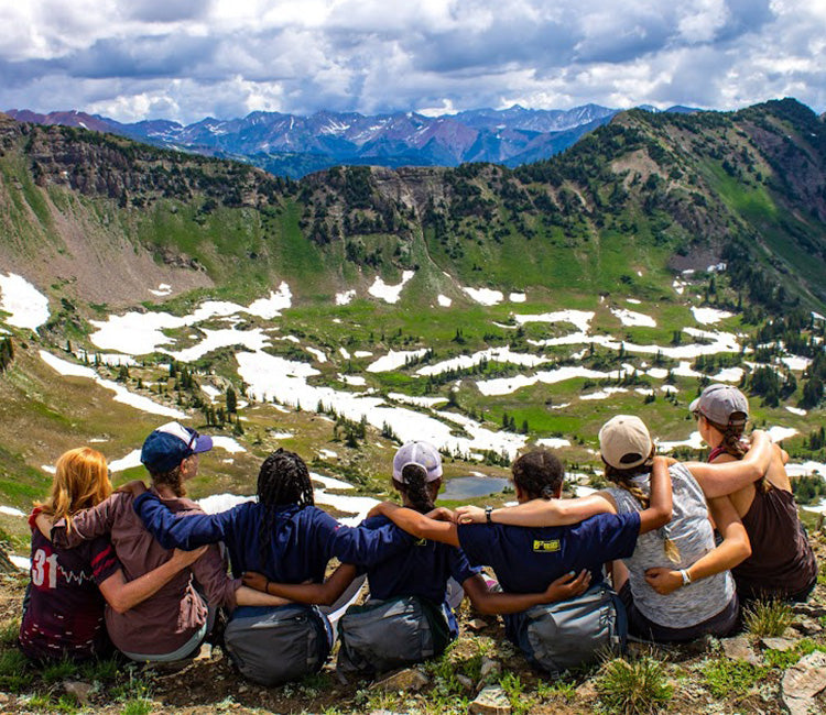 A group of friends in the mountains link arms as they look out on a scenic view