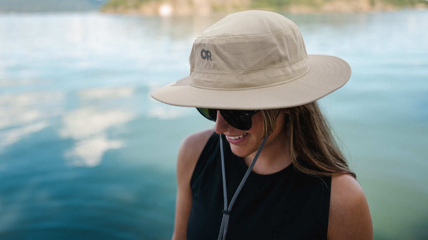 A woman smiles while wearing a sun hat and sunglasses by the water.