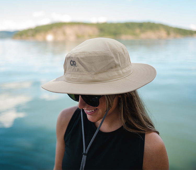 A woman smiles while wearing a sun hat and sunglasses by the water.