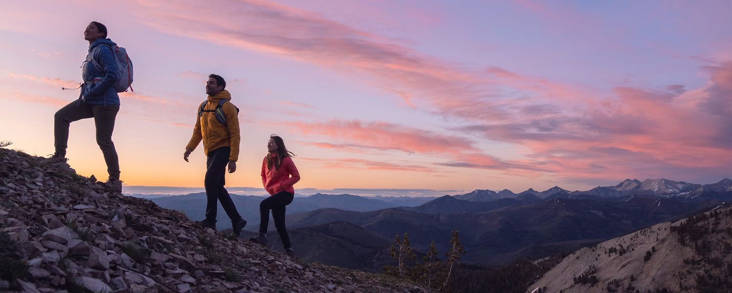 Three friends hike up a rocky trail as the sun sets over the mountains in the background.