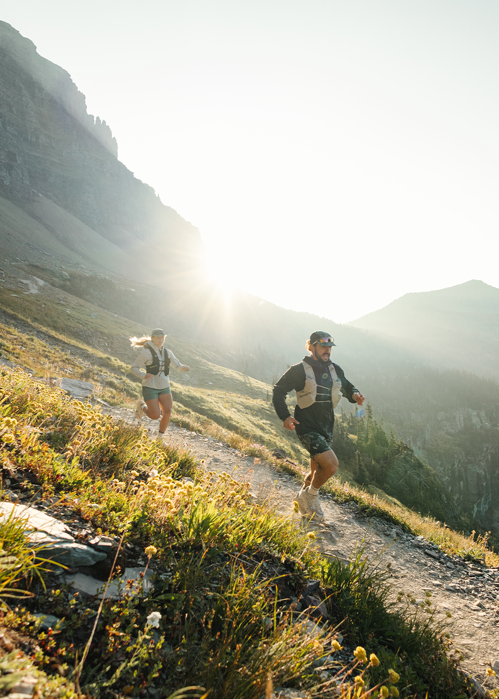 Two people trail running in the mountains with the sun behind wearing Outdoor Research sportswear. 