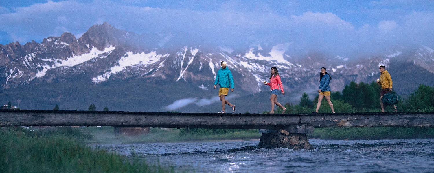 A group of friends walk across a rustic bridge at dusk, with snow-capped mountains in the background.