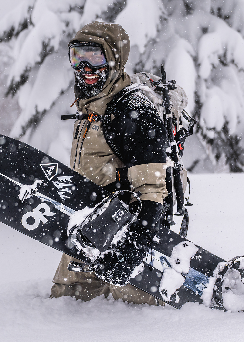 Male hiking through the snow with snowboard wearing Outdoor Research Men's Hemispheres Team GORE-TEX 3l Jacket Gravel/Black. 