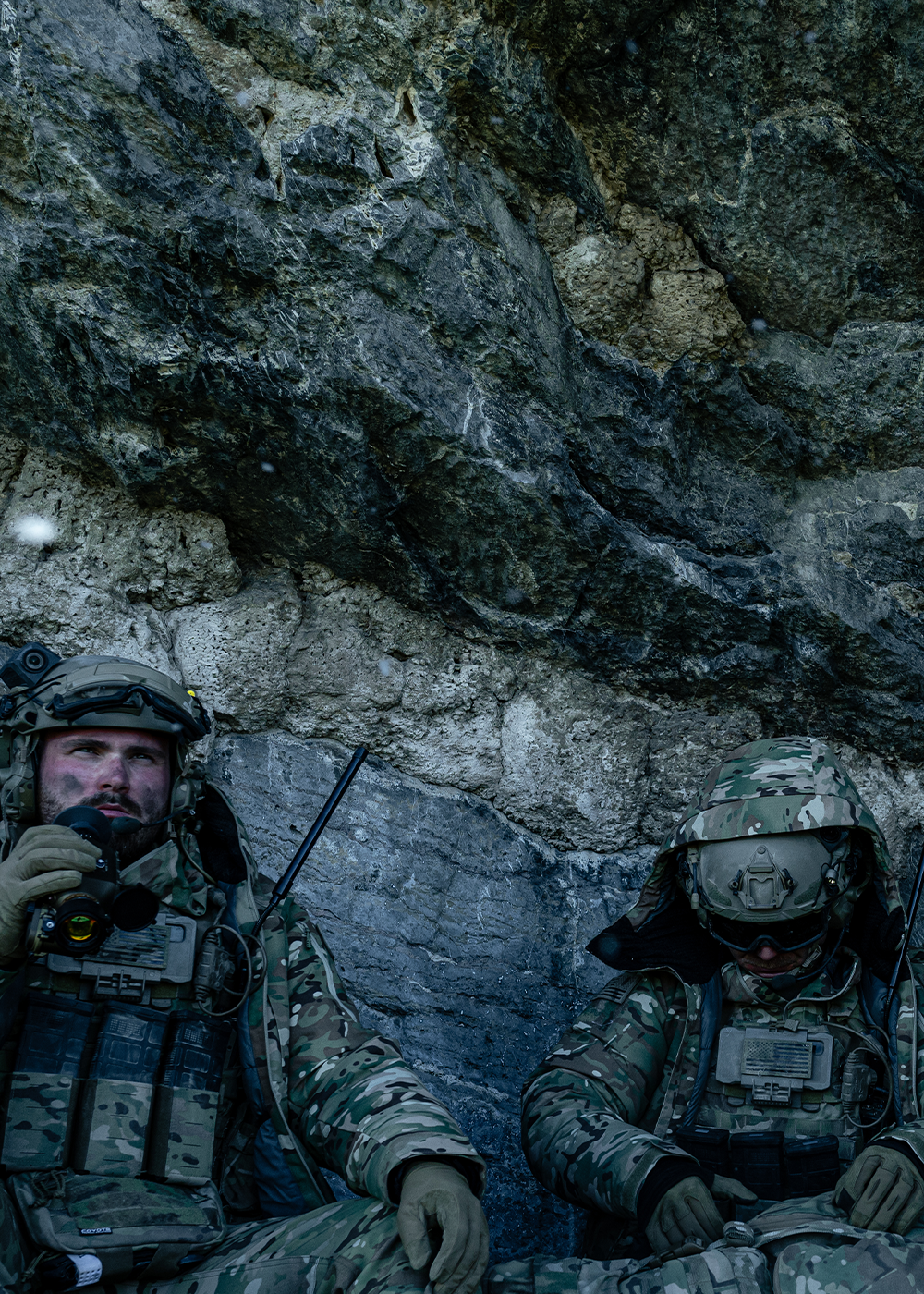 A group of men wear tactical camouflage while leaning up against a rock wall.