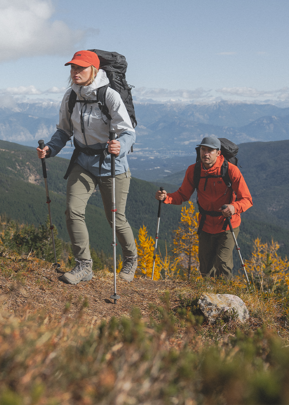 Two hikers on a sunny day wearing Outdoor Research Women's Aspire 3L Jacket Titanium/Ascent Blue, Women's timberline Pant Ranger Green, Mens Ferrosi Hoodie Cardinal. 
