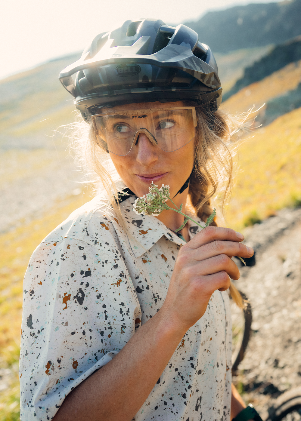 Female holding flowers in her hand in the sun wearing Outdoor Research Women's Astroman Short Sleeve Sun Shirt Gravel Creekside Print.