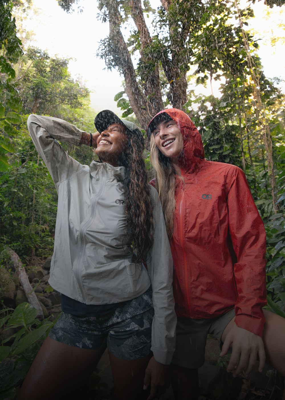 Two females smiling in the rain wearing Outdoor Research Women's Helium UL Jackets Madrone Red and Oyster. 