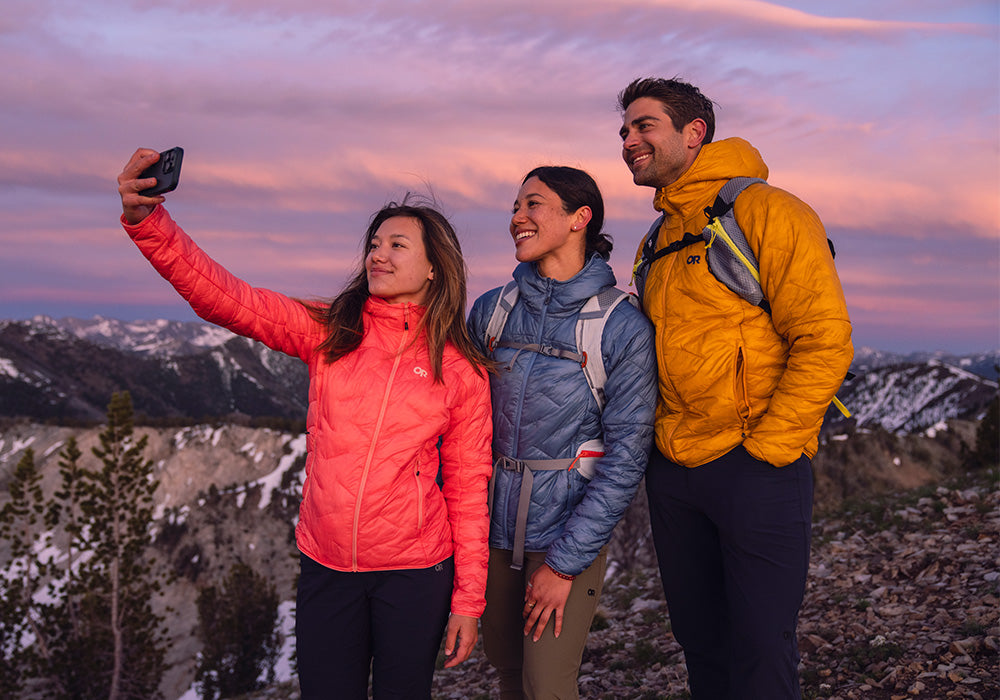 Three friends in colorful jackets take a selfie while standing atop a mountainous overlook.