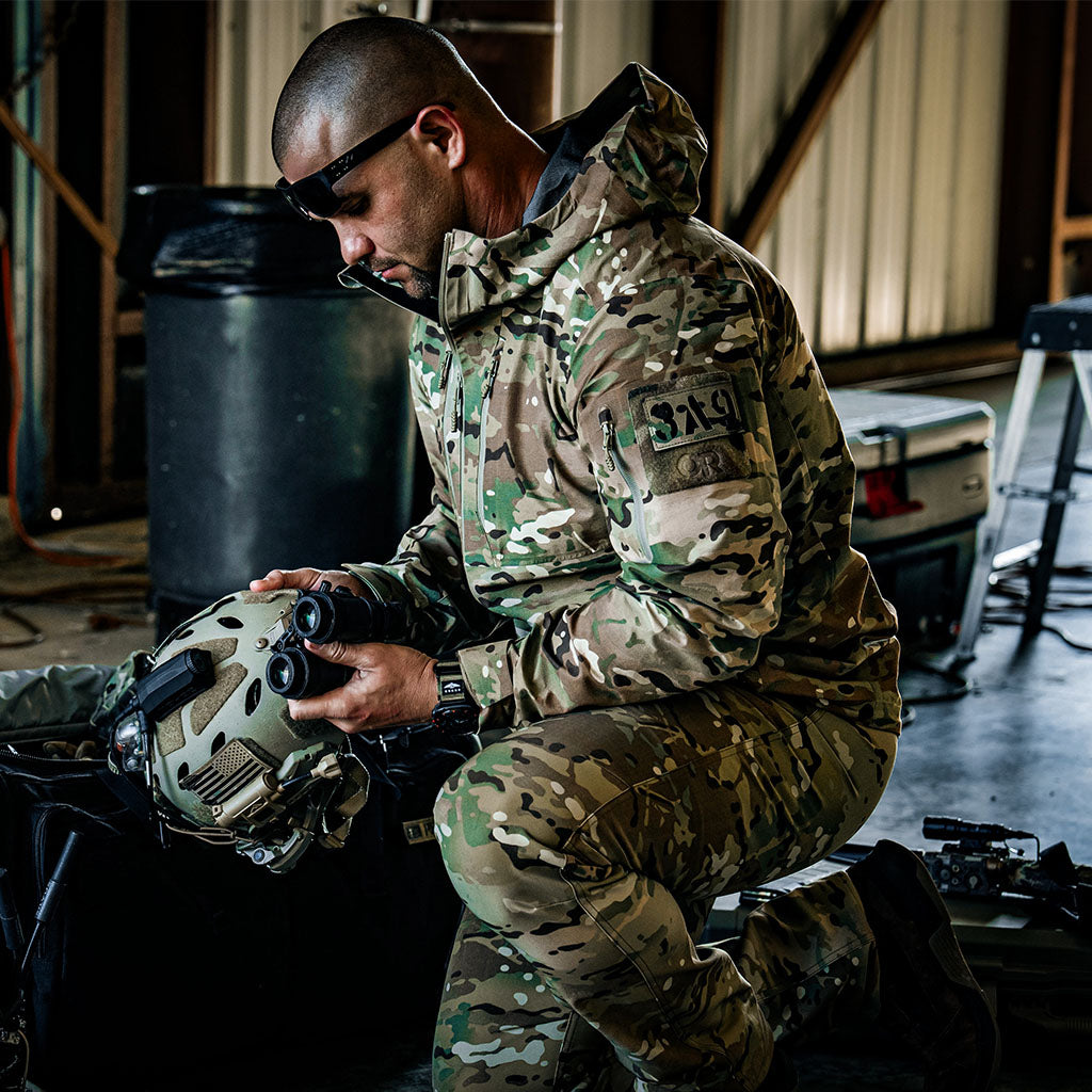 Person in military camouflage gear holding a helmet and binoculars in an indoor setting.